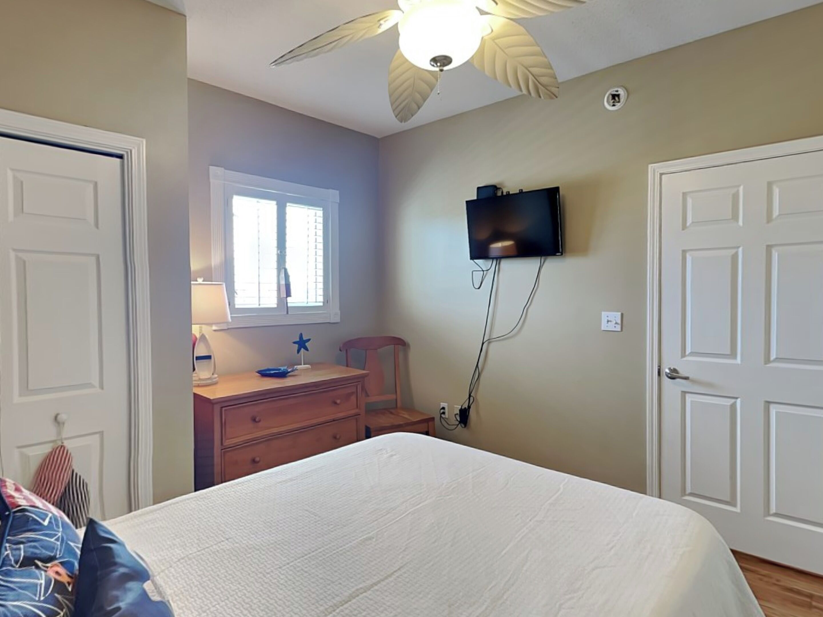 A bedroom with a bed, a dresser, a television mounted on the wall, and a ceiling fan.