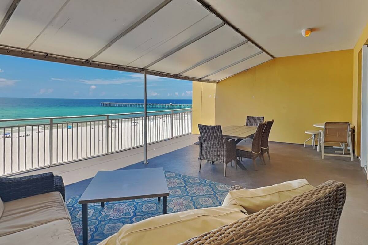 A balcony with a view of the ocean and a pier, furnished with a table, chairs, and a sofa.