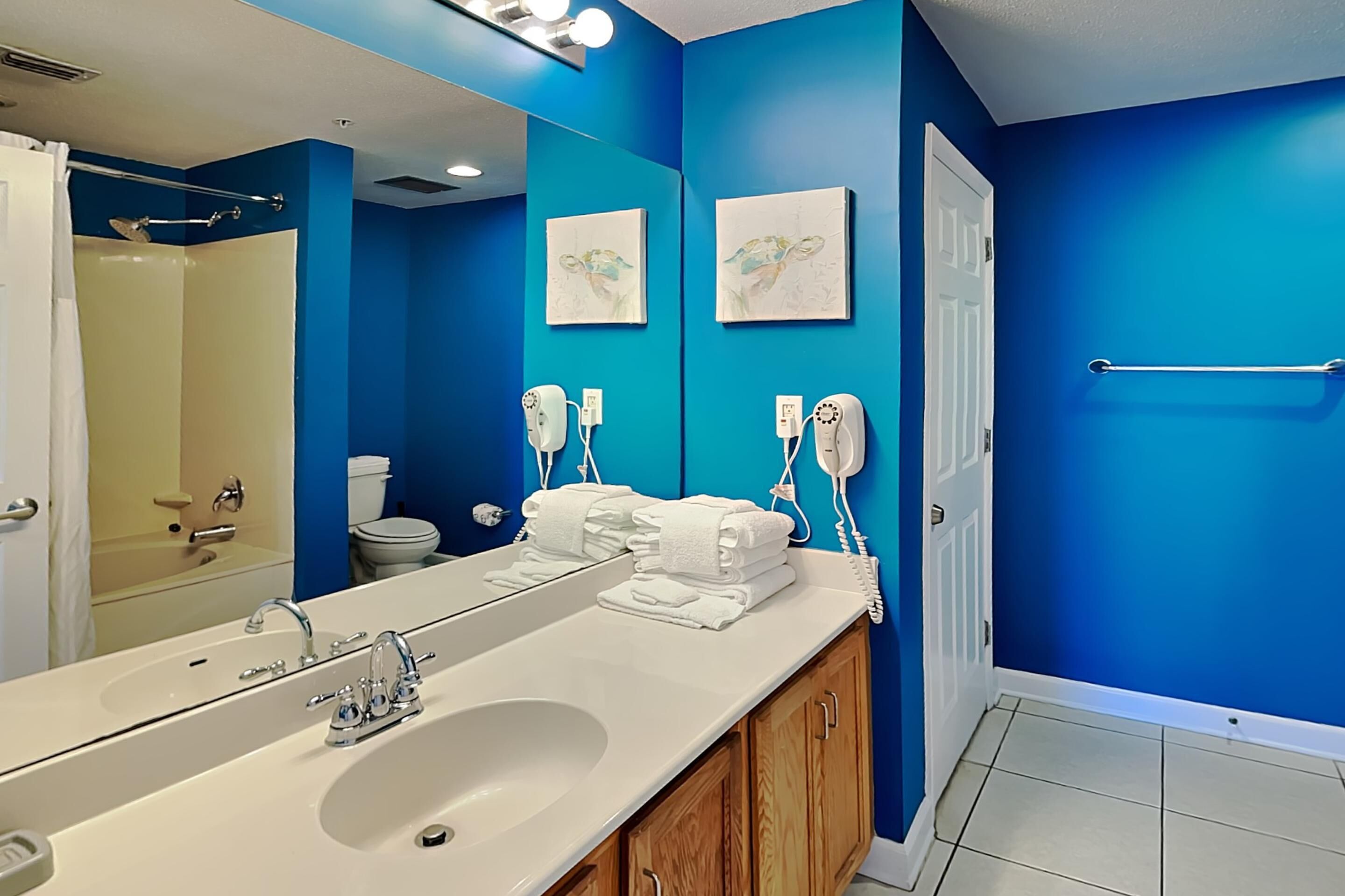 Baño con un espejo grande, lavamanos blanco y paredes azules. Hay dos secamanos y un perchero para toallas.