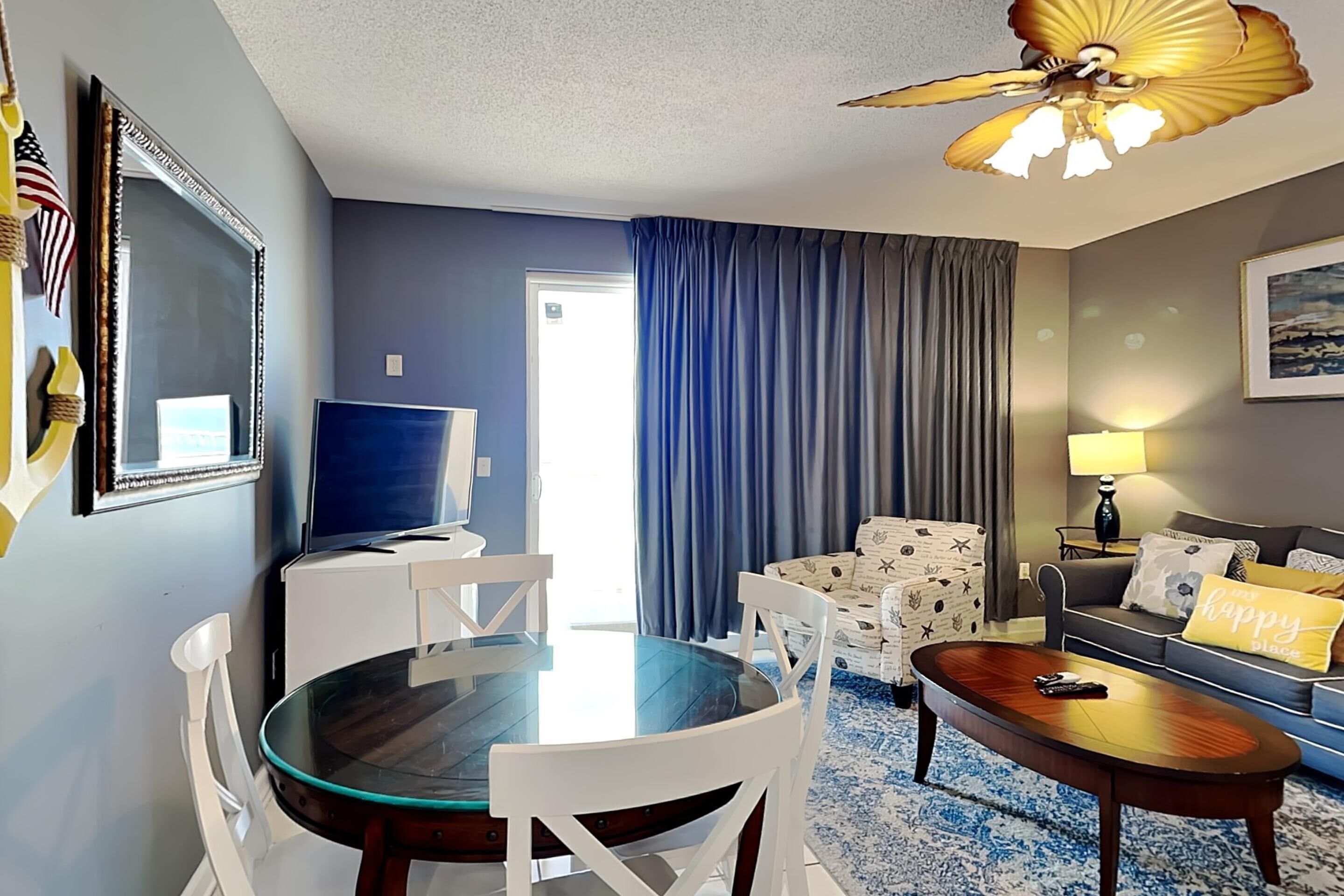 A hotel room with a dining table, chairs, a sofa, a TV, and a ceiling fan.