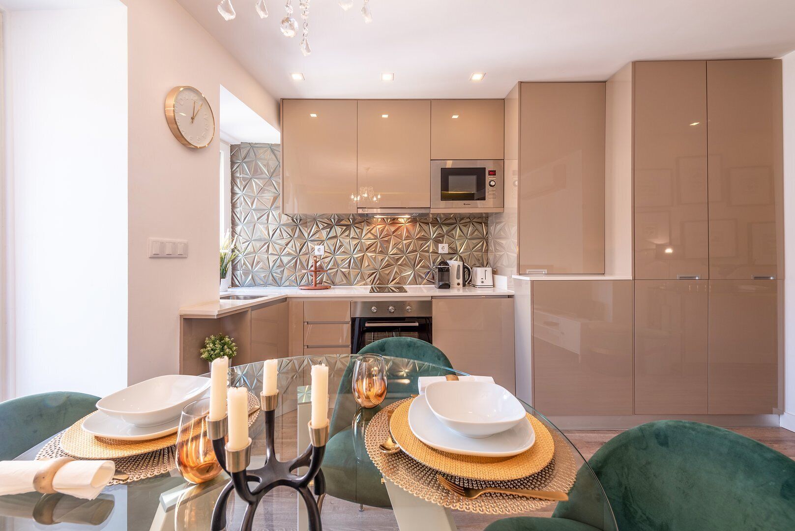 A modern kitchen with a microwave, oven, and a geometric backsplash. There is a dining area with a table set for two, featuring a green chair and a glass table.