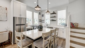 A modern kitchen with white cabinetry, a stainless steel refrigerator, a central island with chairs, pendant lighting, and a view of the city through large windows. - Bright, modern home with shaded porch, AC & on-site golf, pool, beach access (Port Aransas)