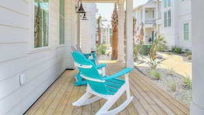A covered porch with two rocking chairs, a wooden deck, and a view of a residential area with palm trees and a building with white siding. - Bright, modern home with shaded porch, AC & on-site golf, pool, beach access (Port Aransas)