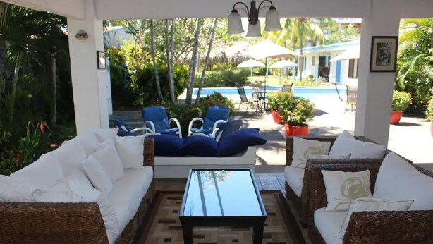 A covered outdoor seating area with wicker furniture and a glass coffee table, overlooking a pool and lush greenery.