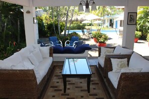 Un espace extérieur aménag é, abrité et doté de meubles en osier et d’une table basse en verre, offrant une vue sur une piscine et une végétation luxuriante.