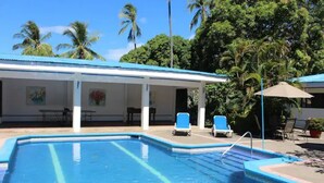 A swimming pool with lounge chairs, a covered patio, and palm trees in the background.