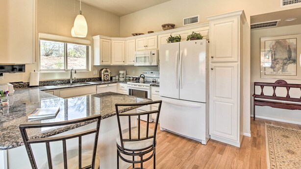 Cucina con mobili bianchi, piano di lavoro in granito, lavello doppio, frigorifero e zona pranzo con due sedie.