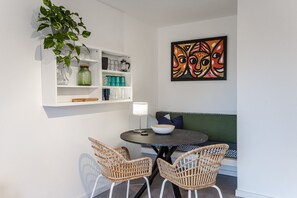 Un coin repas avec des chaises en osier, une table ronde, un saladier, une lampe et une étagère murale où sont disposés des verres et des objets décoratifs.