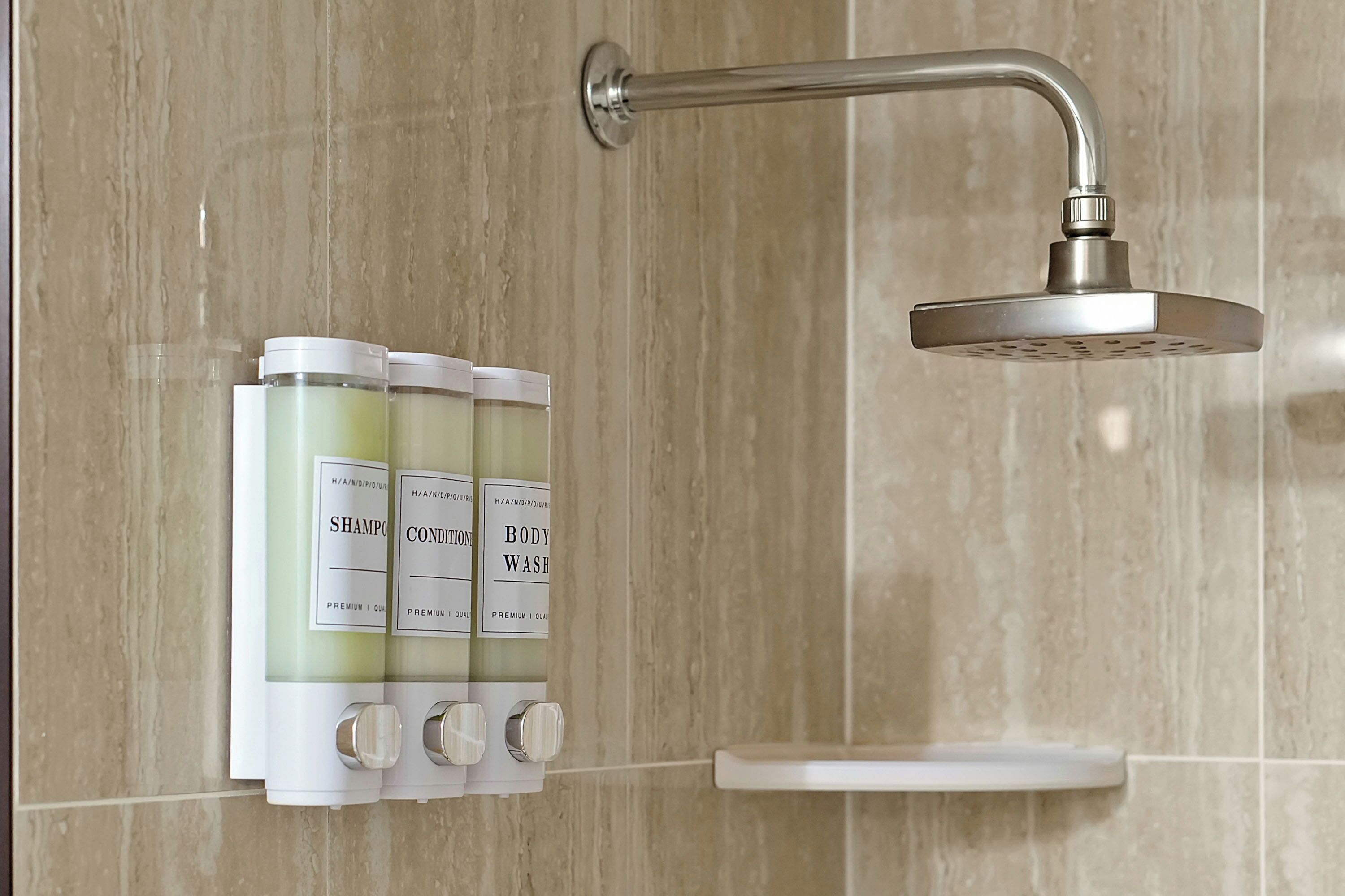 Une salle de bain avec une douche, un distributeur mural contenant trois flacons étiquetés « shampoing », « après-shampoing » et « gel douche », et une étagère blanche.
