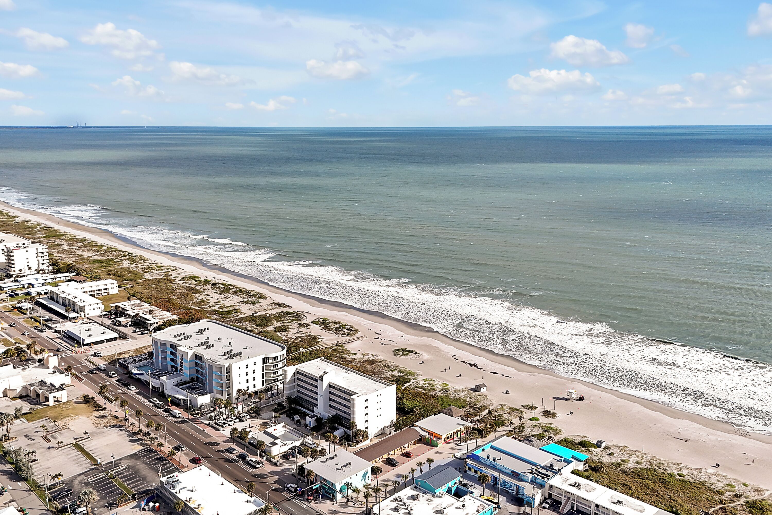 An aerial view of a coastal area with a beach, buildings, and a road.