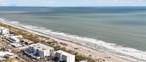 An aerial view of a coastal area with a beach, buildings, and a road.
