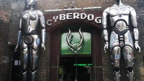 Une entrée d’immeuble avec deux grandes statues de robots métalliques, une façade en briques et une enseigne verte au-dessus de la porte.