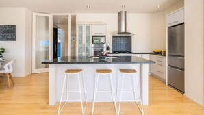 A modern kitchen with a central island, bar stools, stainless steel appliances, and a view into the adjacent dining area. - Hekerua Bay Retreat - Coastal Views (Waiheke Island)