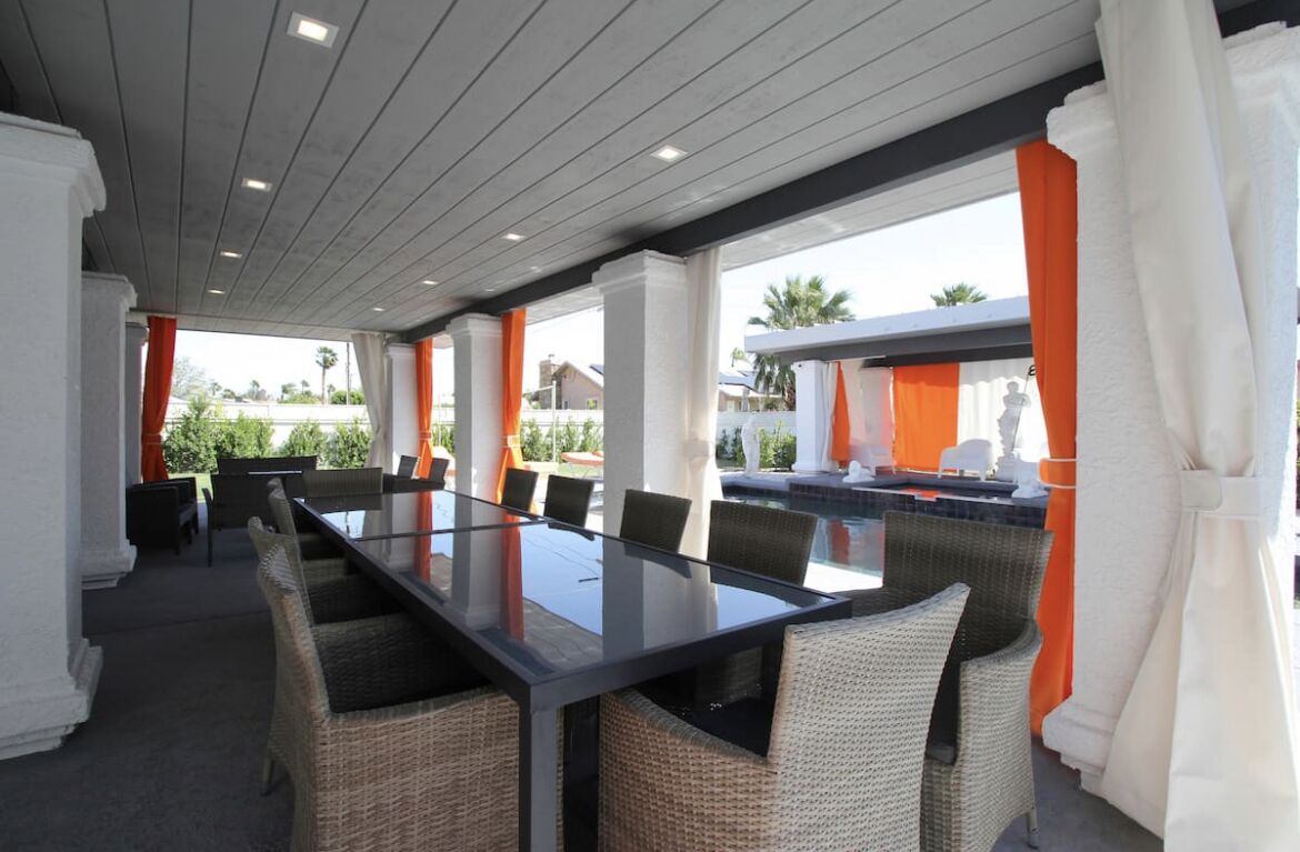 An outdoor dining area with a long table and wicker chairs, surrounded by white columns and orange curtains.