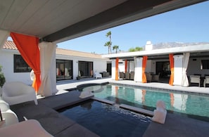 A modern pool area with a clear view of a mountain in the background, a covered seating area, and a white building with large windows.