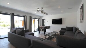 A modern living room with a sofa, coffee table, and a foosball table. There is a TV mounted on the wall and a ceiling fan. - 4 Bedroom Modern Home, Pool+Spa+Guest House! (Palm Springs)