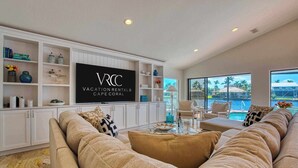 A spacious living room with a large flat-screen TV, a comfortable beige sofa, and a view of a pool through large windows. - VRCC Villa Jelly Key, a boaters dream in paradise (Cape Coral)