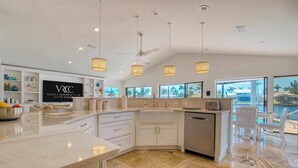 A modern kitchen with white cabinetry, a marble countertop, and a large island. There are pendant lights, a ceiling fan, and a view of the ocean through large windows. - VRCC Villa Jelly Key, a boaters dream in paradise (Cape Coral)