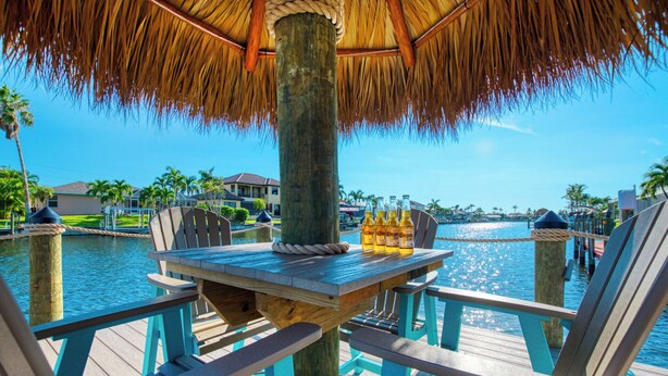 A wooden deck with a thatched roof umbrella, a table with bottles, and chairs. - VRCC Villa Sunny Haven (Cape Coral)