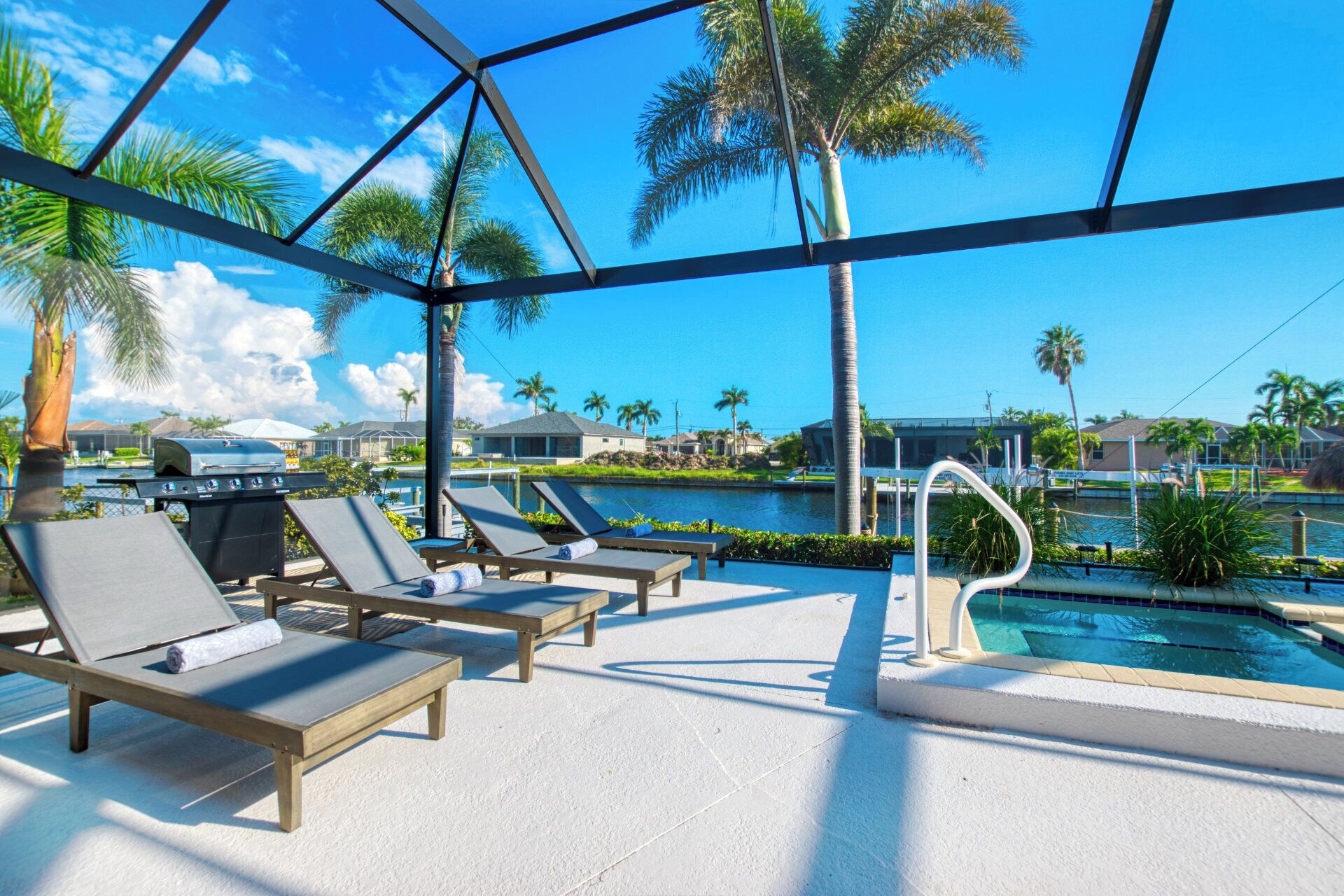 Un espace aménagé au bord de la piscine, avec des chaises longues, un barbecue et une vue sur un plan d’eau et des palmiers.