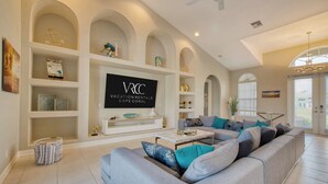 A spacious living room with a grey sectional sofa, a white coffee table, and a TV mounted on the wall displaying 'VRCC Vacation Rentals Cape Coral'. - VRCC Villa Sunny Haven (Cape Coral)