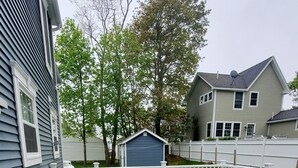Eine Holzterrasse mit weißer Reling, eine blaue Gartenhütte und ein weißer Zaun in einer Wohngegend.