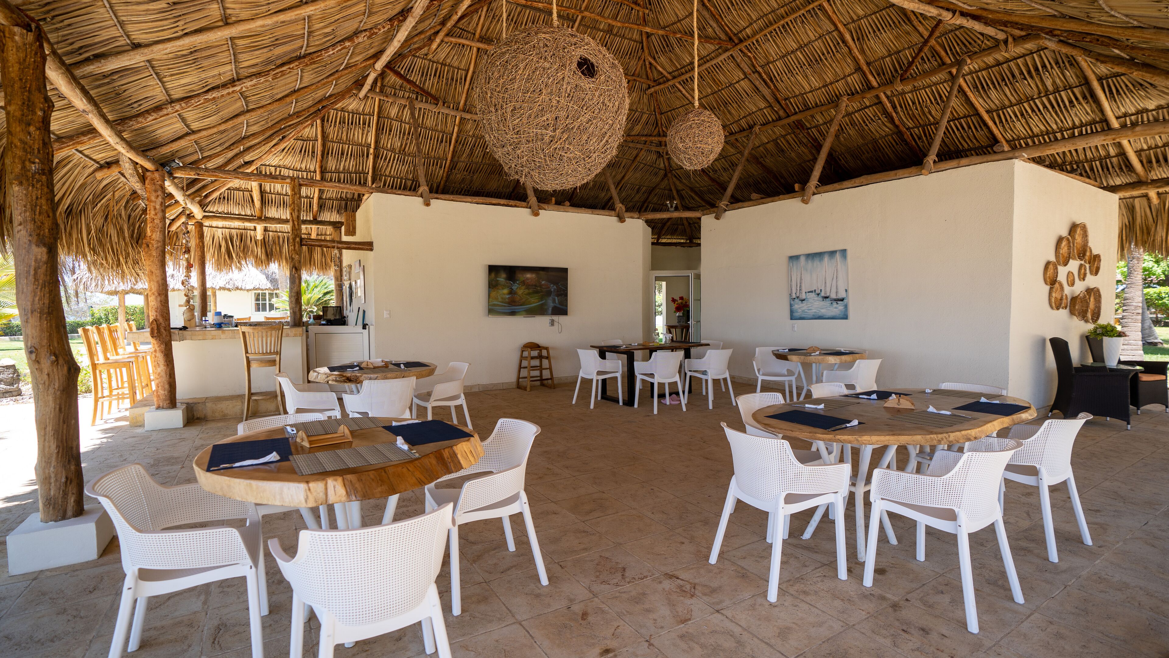 An outdoor dining area with round wooden tables and white wicker chairs, surrounded by a thatched roof structure.