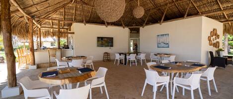 An outdoor dining area with round wooden tables and white wicker chairs, surrounded by a thatched roof structure.