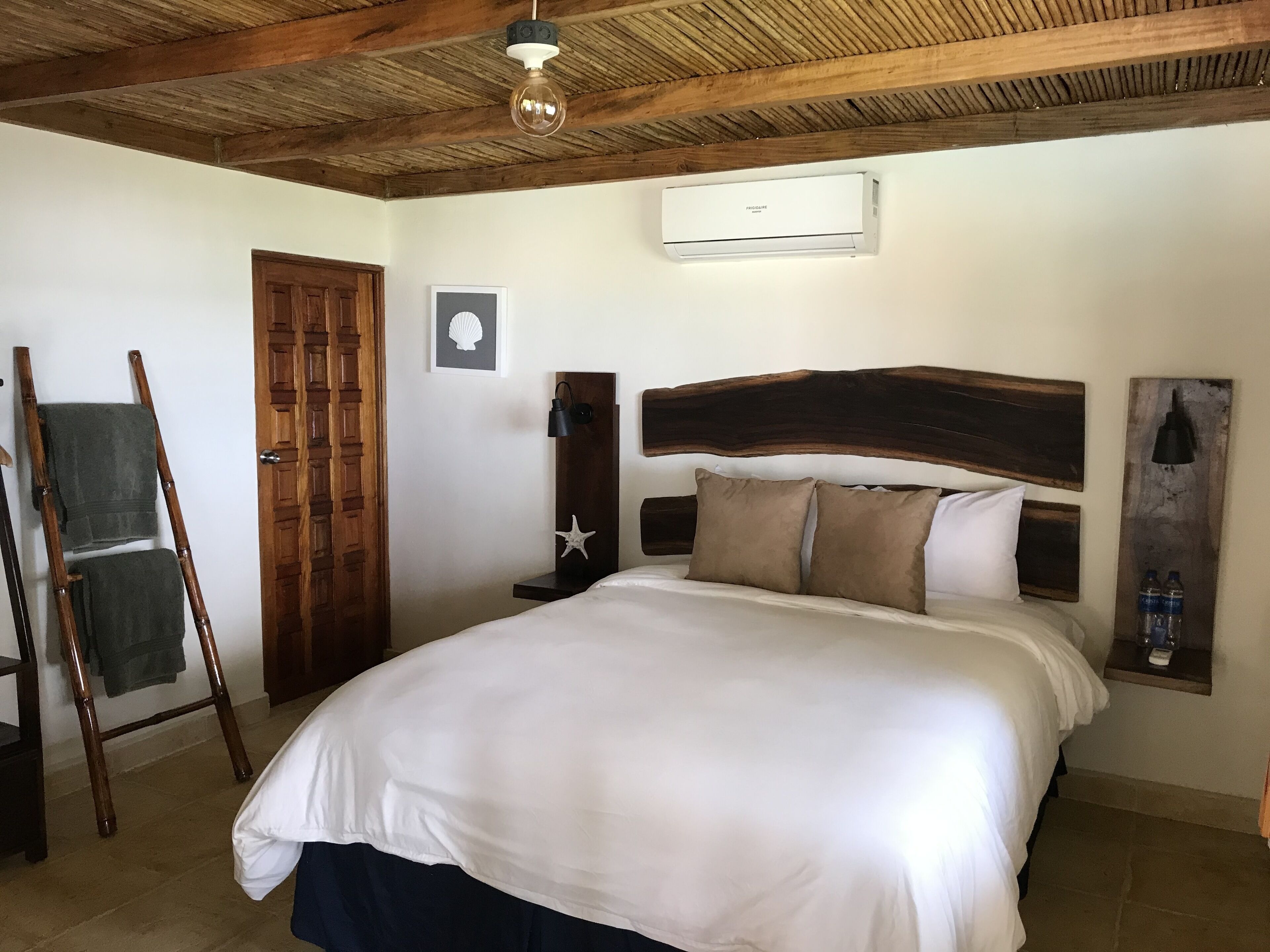 A bedroom with a wooden headboard, a bed with white bedding, a wooden ladder, a door, and a wall-mounted air conditioning unit.