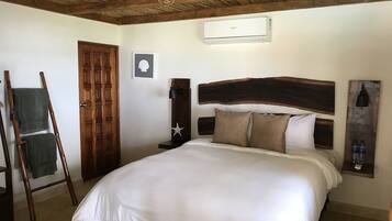A bedroom with a wooden headboard, a bed with white bedding, a wooden ladder, a door, and a wall-mounted air conditioning unit.