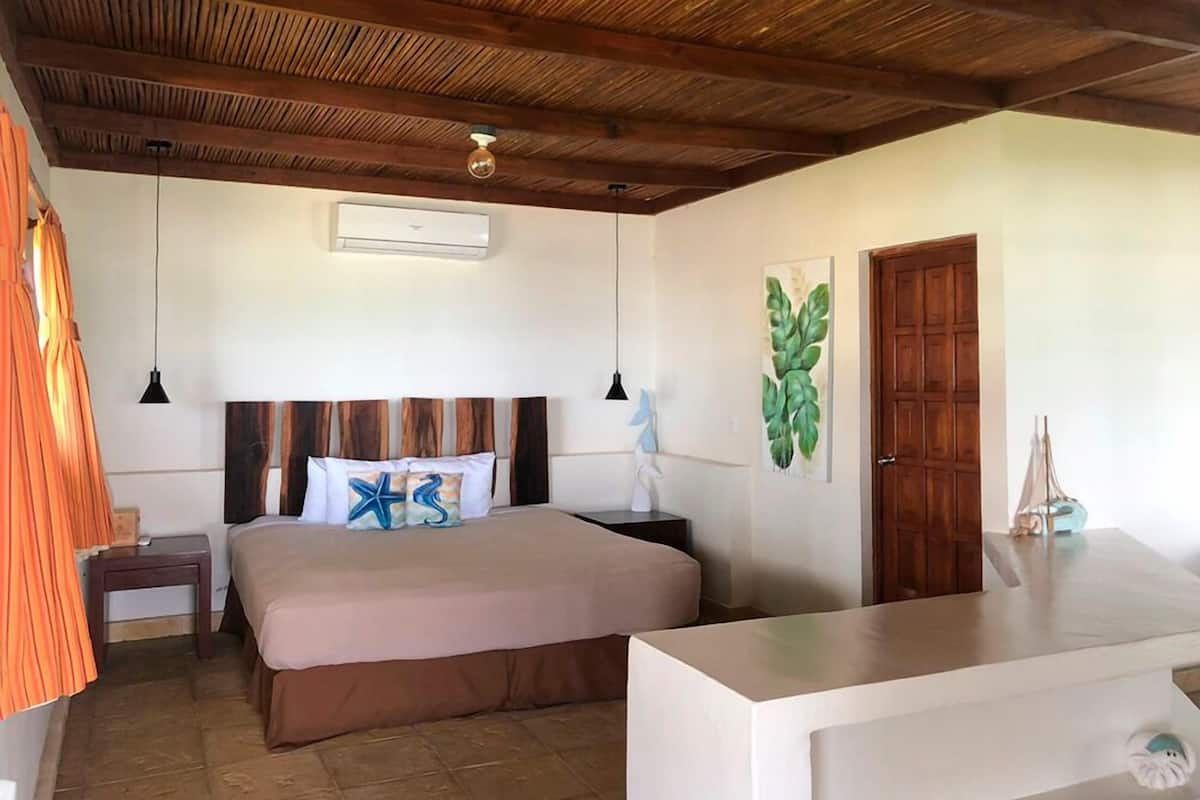 Une chambre d’hôtel avec un plafond en bois, un lit recouvert d’une couvre-lit beige, deux tables de chevet, une petite table et un climatiseur fixé au mur.