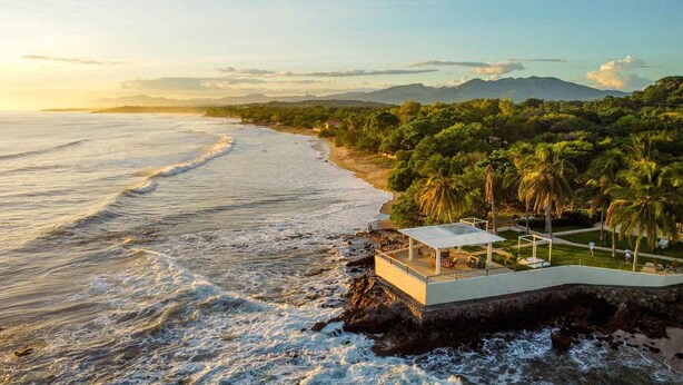 Une propriété située en bord de mer, dotée d’un patio couvert, offrant une vue sur l’océan et s’intégrant dans un paysage verdoyant luxuriant, agrémenté de palmiers.