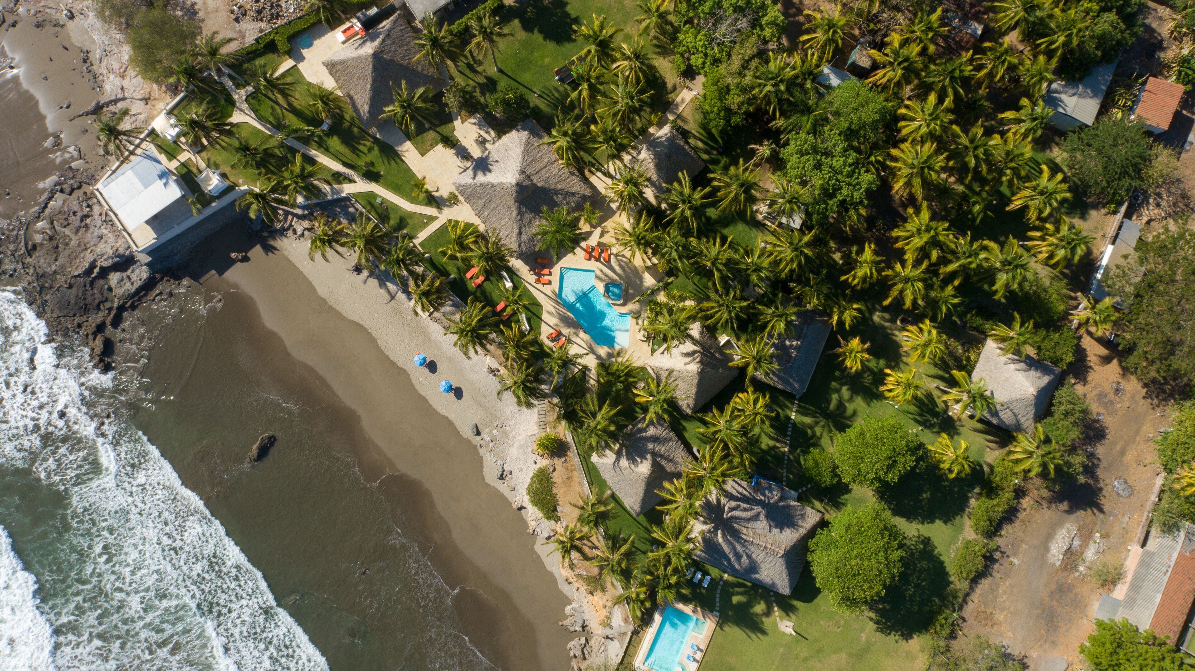 Vue aérienne d’une propriété située sur la côte, avec une piscine, des palmiers et une plage.