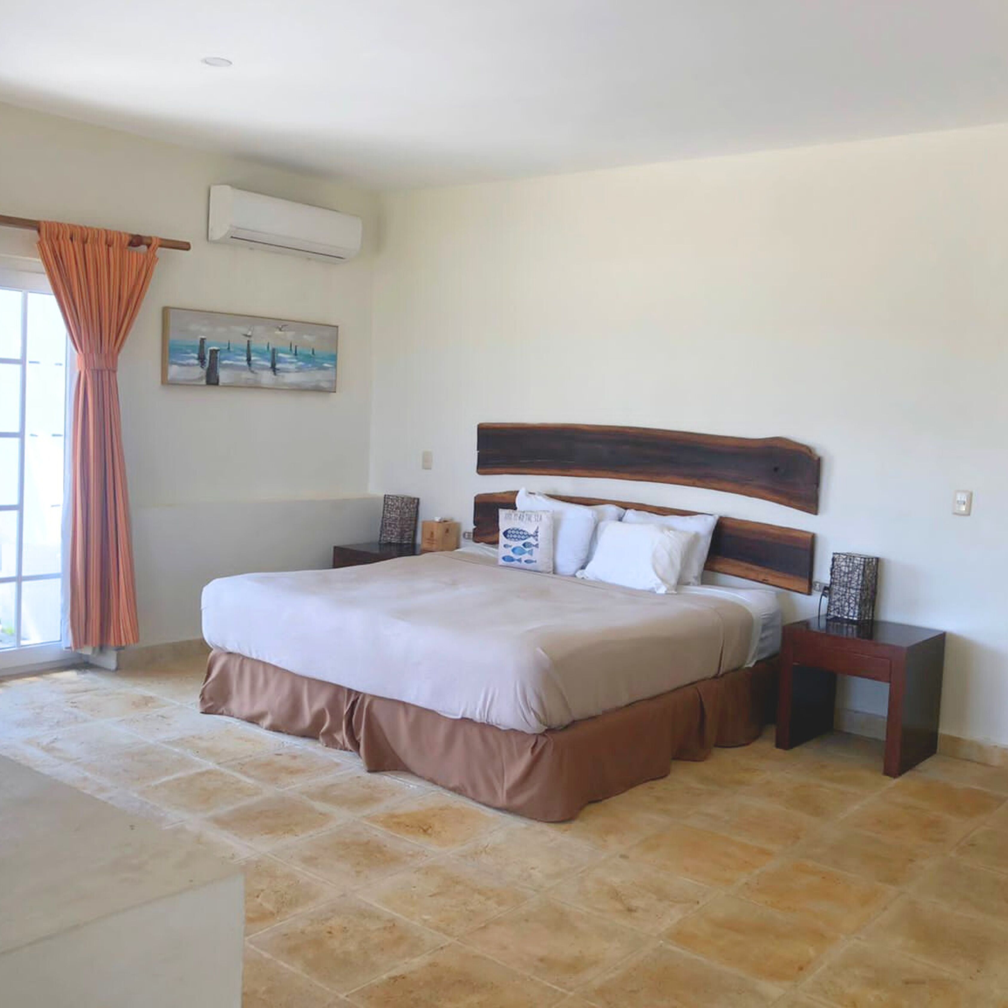 A bedroom with a large bed, a nightstand, a painting on the wall, and an air conditioning unit.
