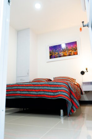 A bedroom with a bed, a white wardrobe, a framed cityscape picture, and a small white shelf.