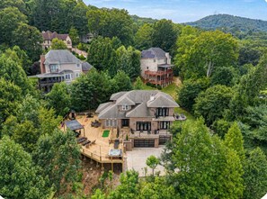 A large house with a deck, surrounded by dense trees and hills.