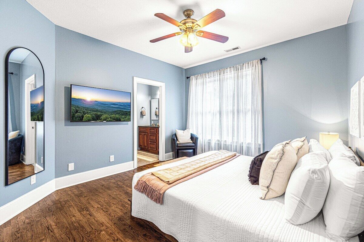 Un dormitorio con una cama, ventilador de techo, un ventanal con cortinas, un espejo y un cuadro en la pared.