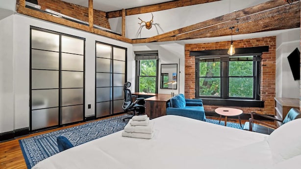 A modern hotel room with exposed brick walls, a large bed with white linens, a desk, a blue sofa, and a large window offering a view of greenery. - Sleeps 14 · Historic 4BR · Steps to Shaw Metro (Washington)