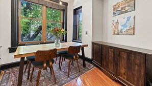 A dining area with a wooden table, four chairs, a vase with flowers, and a large window with a view of trees. - Sleeps 14 · Historic 4BR · Steps to Shaw Metro (Washington)