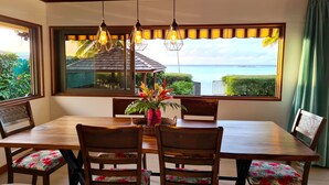 Un comedor con una mesa y sillas de madera, un jarrón con flores, y vista al mar a través de amplios ventanales.