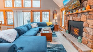 A spacious living room with a large sectional sofa, a fireplace, and a television mounted on a stone wall. - Hot Tub, Sauna & Game Room, Sleeps 8, Pet OK - Near Camelback (Tannersville)