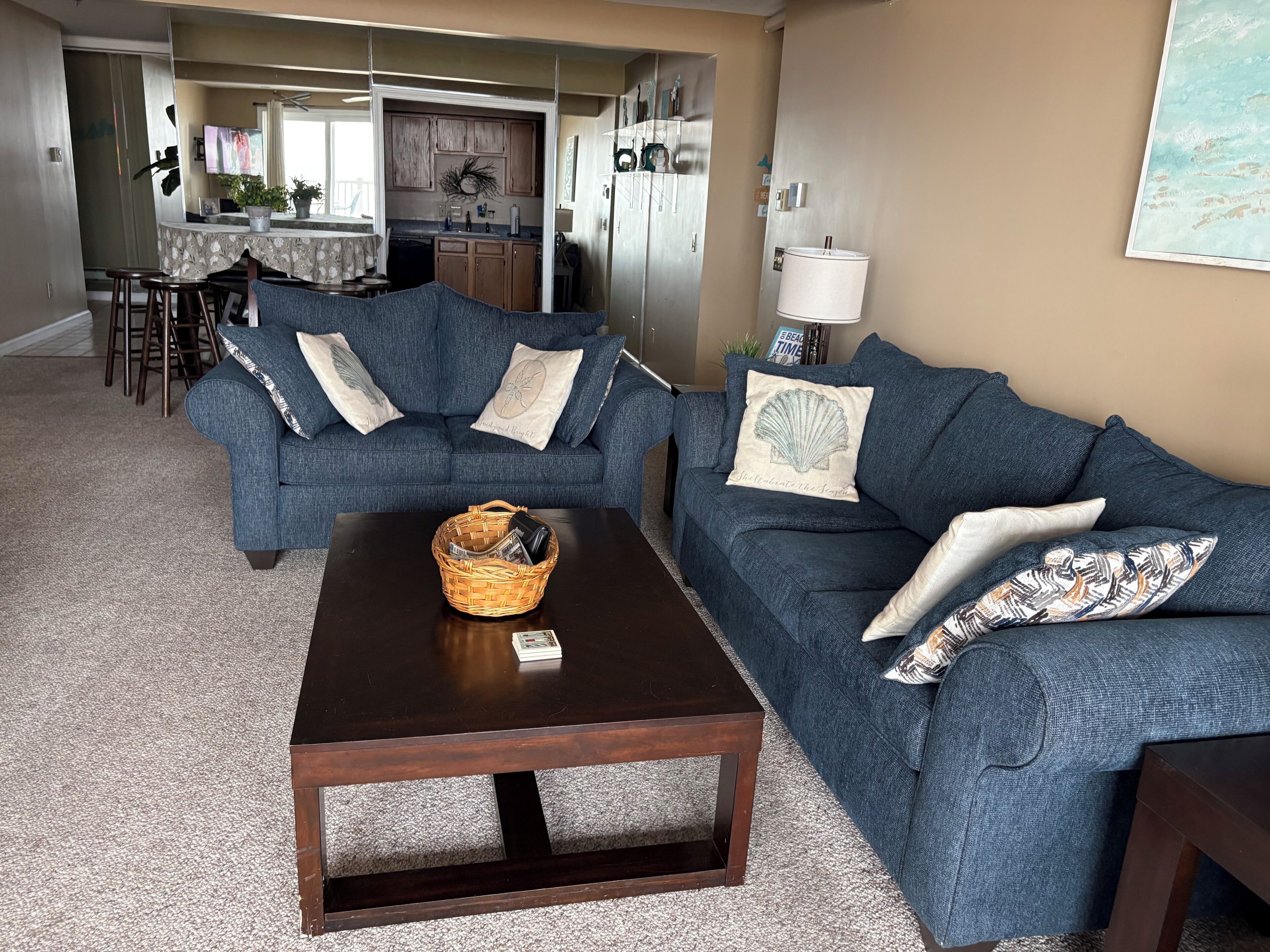 Ein Wohnzimmer mit einem dunklen Holztisch, zwei blauen Sofas mit Zierkissen, einer Küche im Hintergrund und einem Essbereich mit Stühlen.