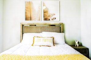 Een net opgemaakt bed met een witte deken en een geel gestreept dekbed, een kussen met de tekst 'STAY AWAY' en een klein beeldje op het nachtkastje.