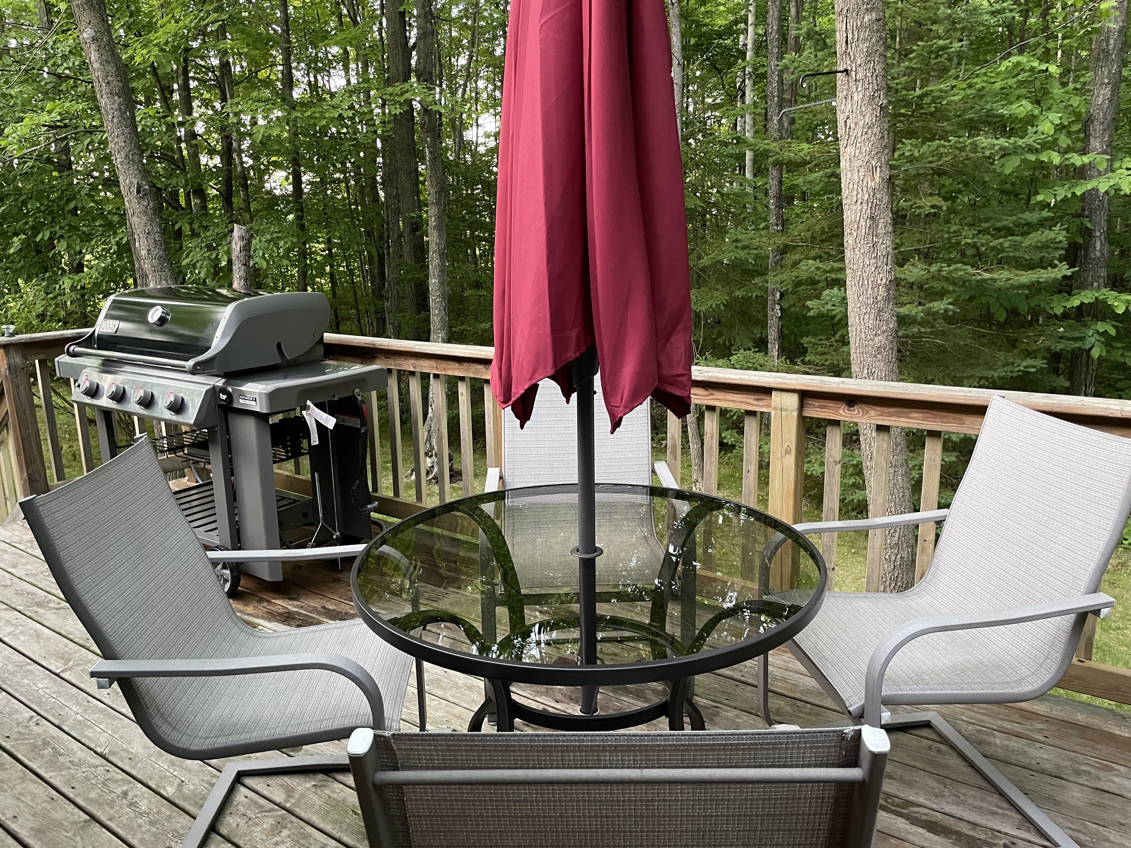 A deck with a glass table, two chairs, a grill, and a red umbrella.