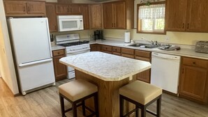 A kitchen with wooden cabinets, a white refrigerator, a microwave, a stove, a sink, and a marble countertop with two stools.