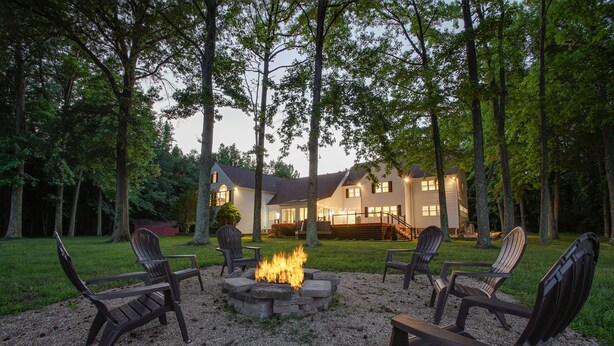 Un foyer est entouré de chaises Adirondack dans un espace boisé, avec une maison en arrière-plan.