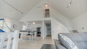 Un vaste espace de vie aménagé dans un style industriel, avec un plafond haut, un escalier menant à un niveau supérieur et une cuisine située au fond de la pièce.