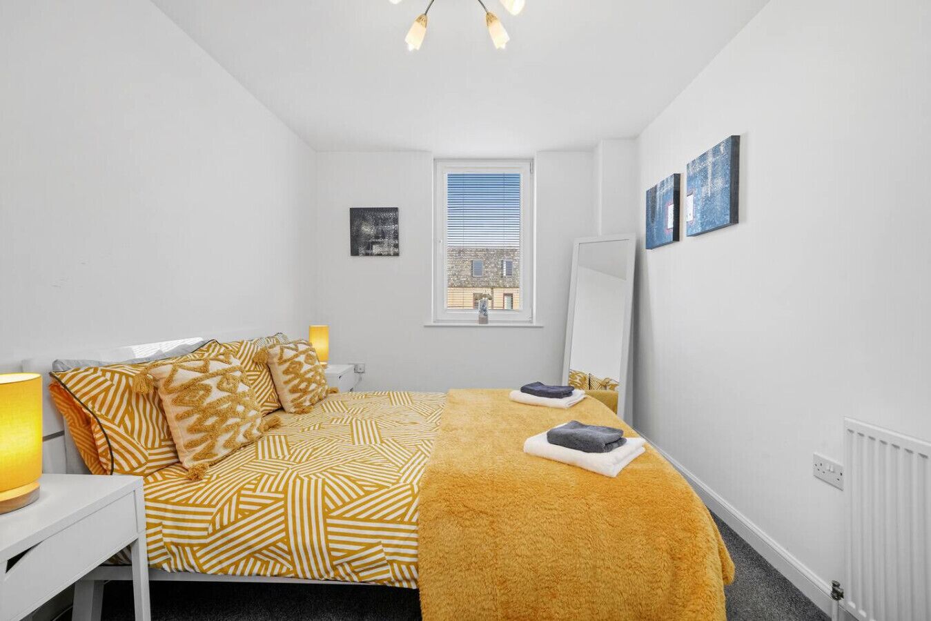 Un dormitorio con cama, mesita de noche, lámpara, ventana y una obra de arte en la pared.