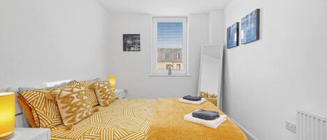 Un dormitorio con cama, mesita de noche, lámpara, ventana y una obra de arte en la pared.