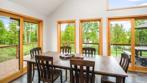Un comedor con mesa y sillas de madera, ventanas amplias y vistas a una terraza y árboles.